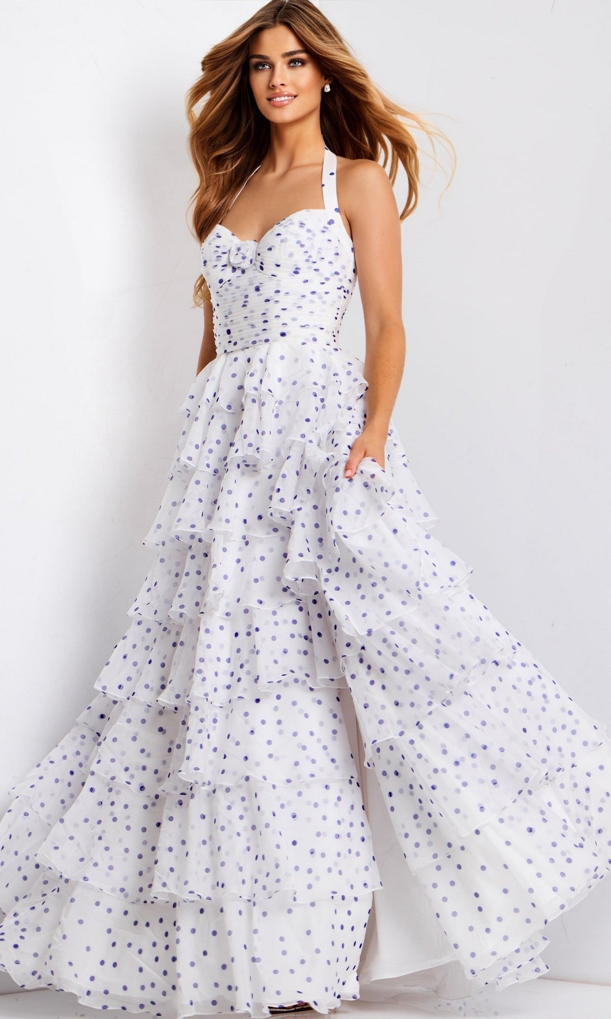 A woman stands in front of a plain background wearing the Jovani 49776 Long Prom Dress, a white floor-length gown with purple polka dots and a sweetheart halter neckline.